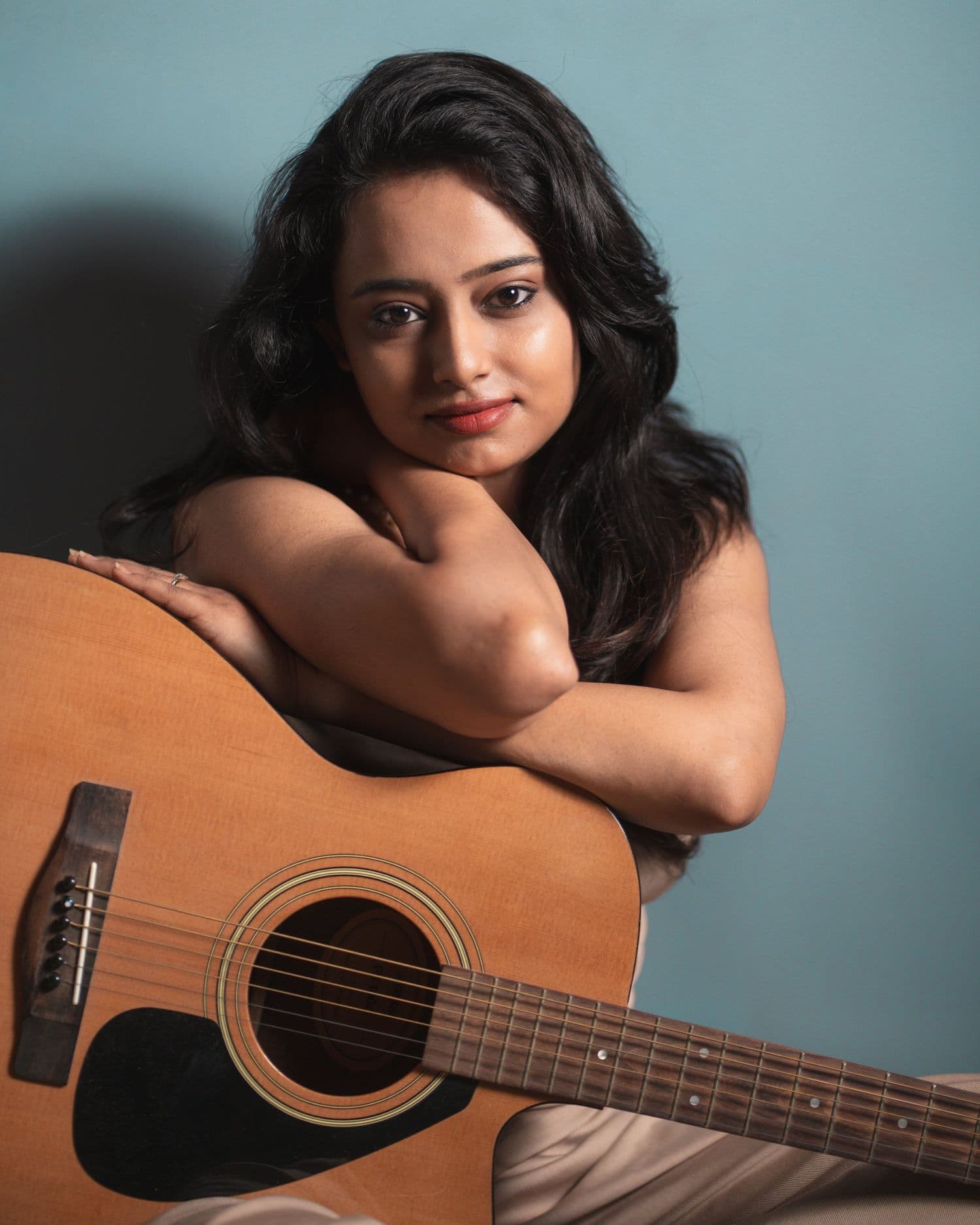 Close portrait of Ankita Barwad leaning on the body of an acoustic guitar against a teal wall.