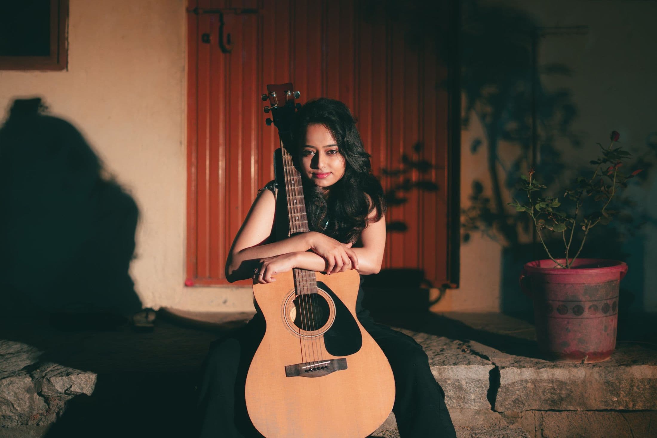 Ankita Barwad sitting on a stone step in warm evening light, resting her arms on an acoustic guitar in front of a rust-red door.