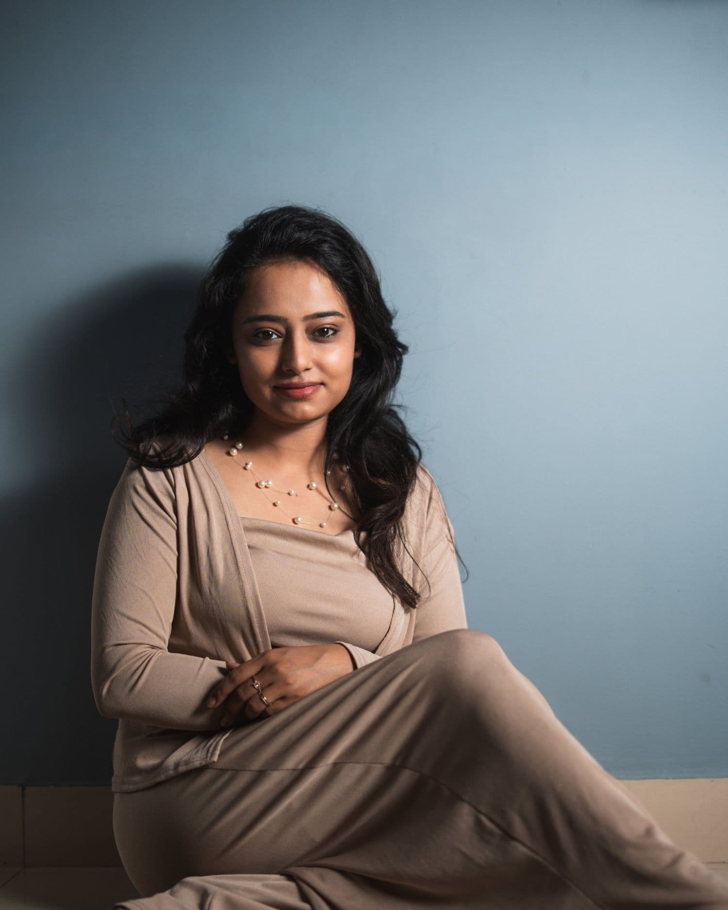 Portrait of Ankita Barwad sitting against a blue wall in a soft beige outfit and pearl necklace, smiling gently.